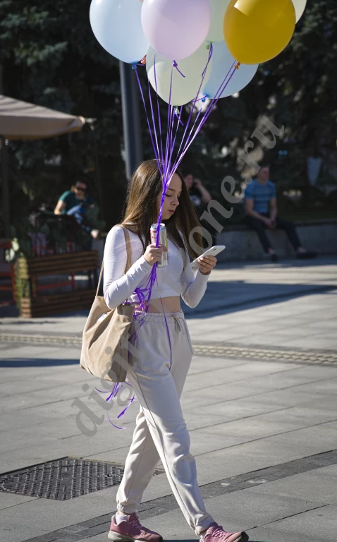 По центральной улице города, залитой солнечным светом, идет девушка со связкой цветных воздушных ...