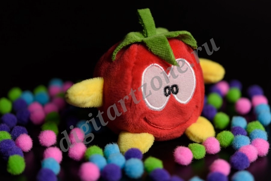 Композиция из мягкой игрушки в виде помидора и цветными помпонами вокруг неё на чёрном фоне.